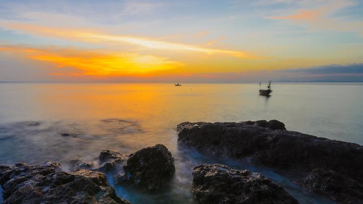 Sonnenuntergang über ruhigem Wasser, Felsen im Vordergrund und ein Boot entfernt auf dem Meer.